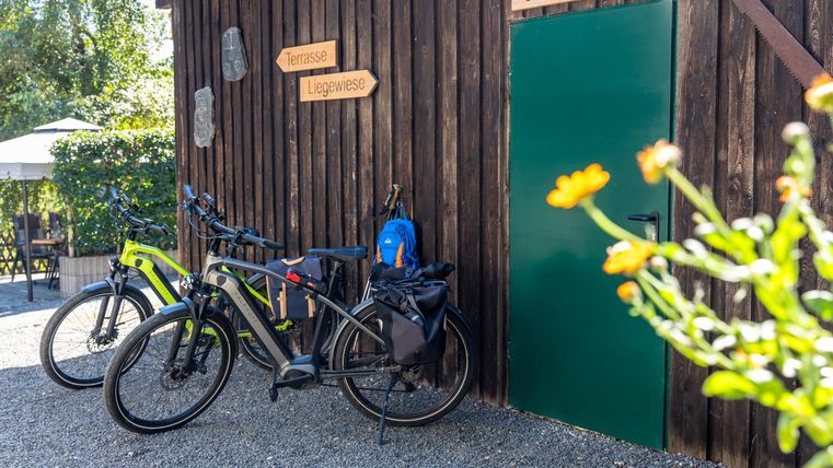 Een fietsgarage van hout met twee fietsen en een blauwe rugzak. Op de achtergrond zijn planten en een tafel in de buitenlucht te zien.