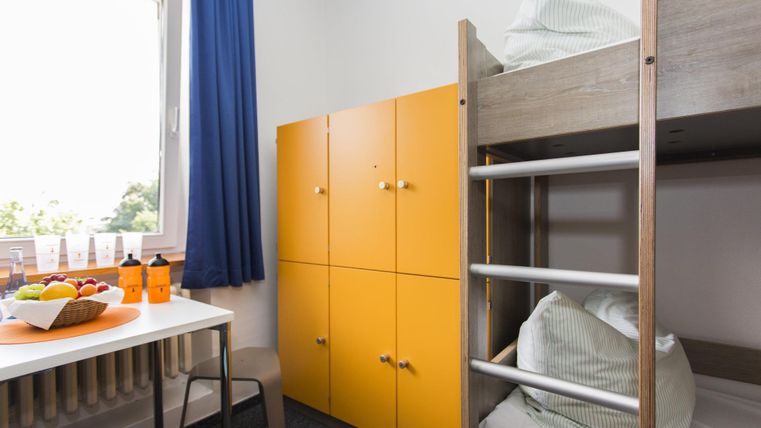 A simple room with a loft bed and orange cabinets. On the table, there is a bowl of fruit and drinks.