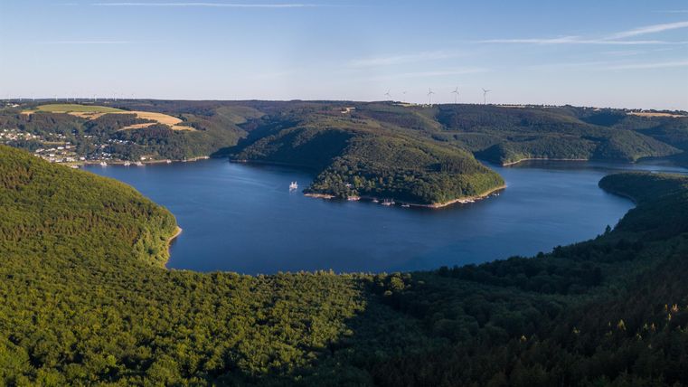 A picturesque landscape with a large lake, surrounded by hills and dense forest. The sky is clear and blue, perfect for outdoor activities.