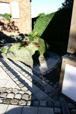 A paved path runs through a small garden with green plants. In the foreground, the shadow of a person is visible.