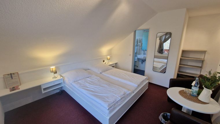 A cozy bedroom with a double bed and modern furniture. In the background, a bathroom and a small living area are visible.