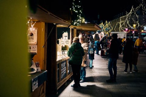 Ein belebter Weihnachtsmarkt bei Nacht mit festlicher Beleuchtung. Menschen stöbern an Holzständen mit Waren.