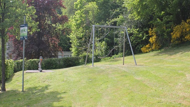 A quiet park with a swing and lush green grass. In the background, there are trees and a bus stop.