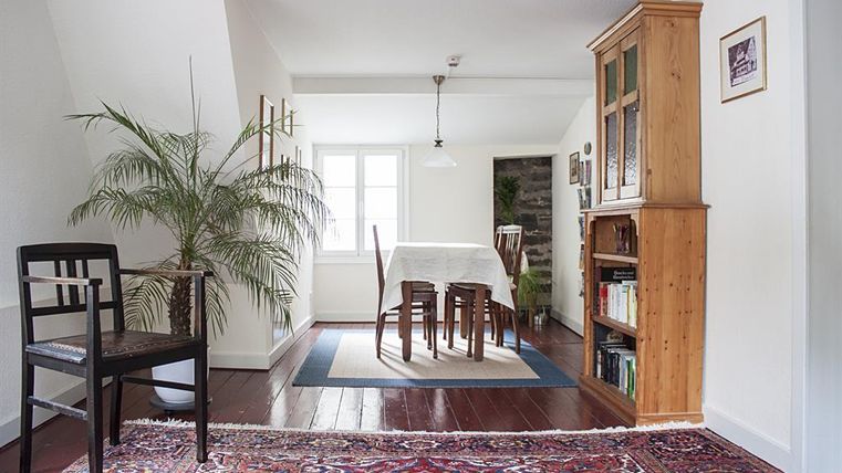 Een heldere, uitnodigende eetruimte met een tafel en stoelen. Een grote plant en een boekenplank geven de ruimte een gezellige sfeer.
