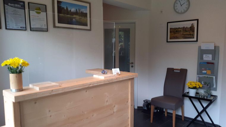 Ein Empfangsbereich mit einem Holztresen und einer Pflanze auf dem Tisch. An der Wand hängen Bilder und Urkunden, und es gibt einen Stuhl mit Blumen.