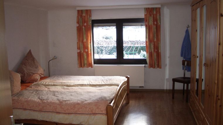 A simple bedroom with a large bed and a wooden wardrobe. The room has a window with curtains and a chair in the background.