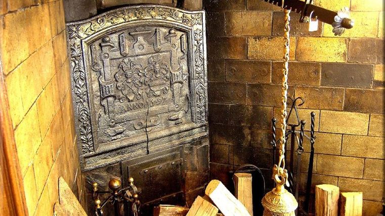 Ein traditioneller Kamin mit einer kunstvollen Metallverkleidung und Holzscheiten im Vordergrund. Der Raum hat eine gemütliche Atmosphäre und rustikale Details.