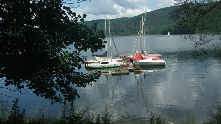 A tranquil lake landscape with several sailboats. In the background, gentle hills and a blue sky are visible.