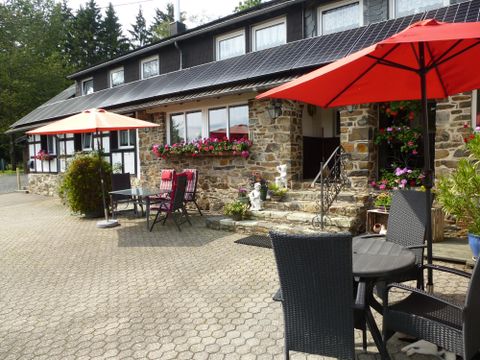 Een gezellig restaurant met een stenen gevel, omringd door kleurrijke bloemen. Op het plein staan tafels met rode parasols.