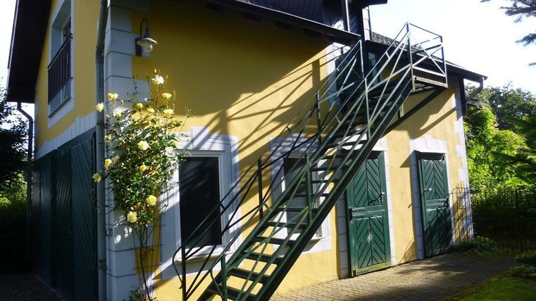 A charming house with a yellow facade and green doors. A staircase leads to the upper floor, surrounded by a green landscape.