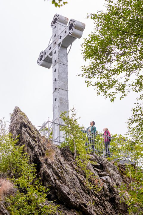 A large, modern cross stands on a rock in the forest. Two people are standing near the cross and enjoying the view.