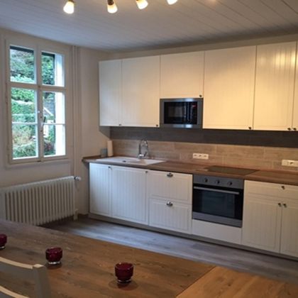 A modern kitchen with white cabinets and a wooden countertop. A dining table with red candles stands in the foreground and a window lets in plenty of natural light.