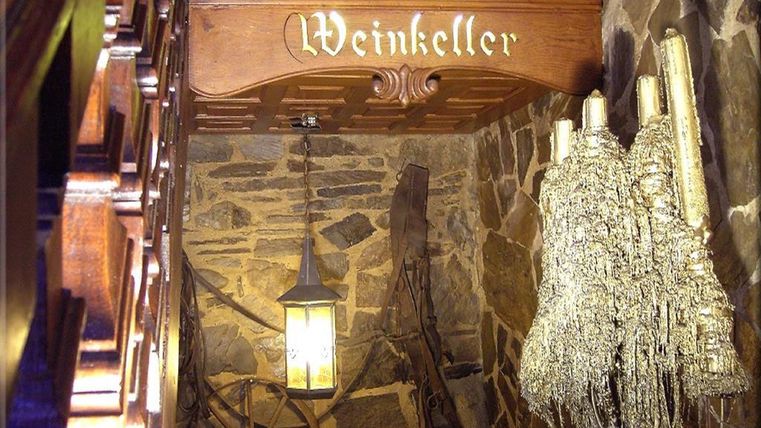 Ein Weinkeller mit rustikalen Steinwänden und einer hölzernen Decke. Eine hängende Laterne und Dekorationen verleihen dem Raum Gemütlichkeit.