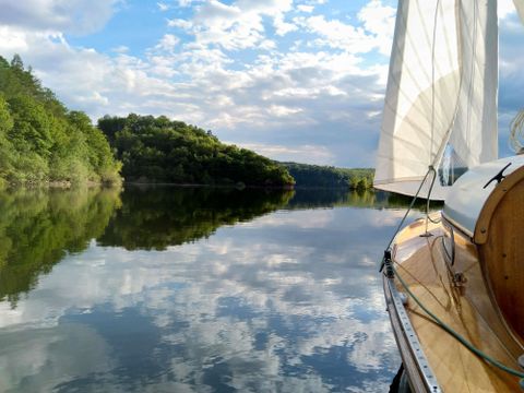 A sailboat is sailing on a calm body of water, surrounded by green hills. The sky is clearly reflected in the water.