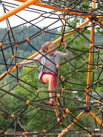 A child is climbing on a climbing frame made of ropes. In the background, trees and a green landscape can be seen.