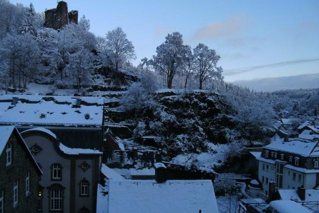 A winter landscape with snow-covered trees and a hill in the background. On the hill stands an old castle, surrounded by quiet houses.