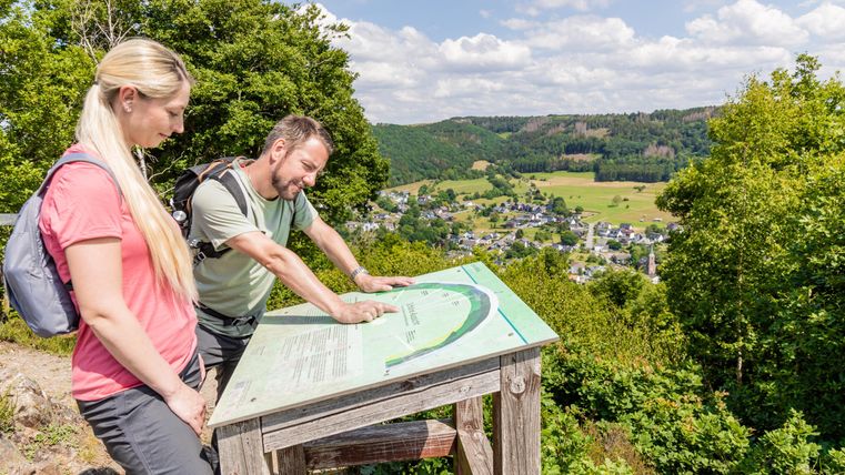 Zwei Wanderer betrachten eine Karte auf einem Aussichtspunkt. Im Hintergrund ist eine schöne Landschaft mit Hügeln und einem kleinen Dorf zu sehen.