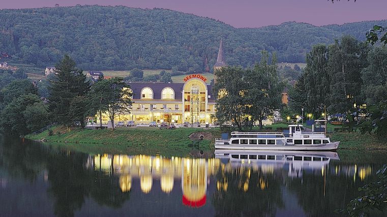 A picturesque view of a building by the river with gentle hills in the background. The light from the building is reflected on the water.