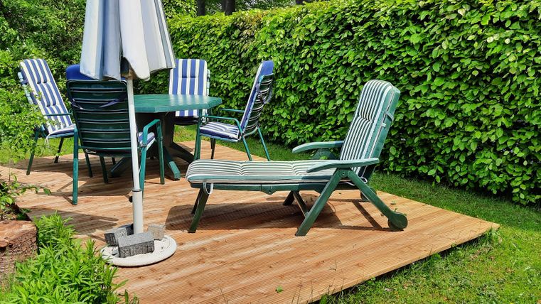 A cozy terrace with a table and wooden chairs. In the background, a green hedge is visible.