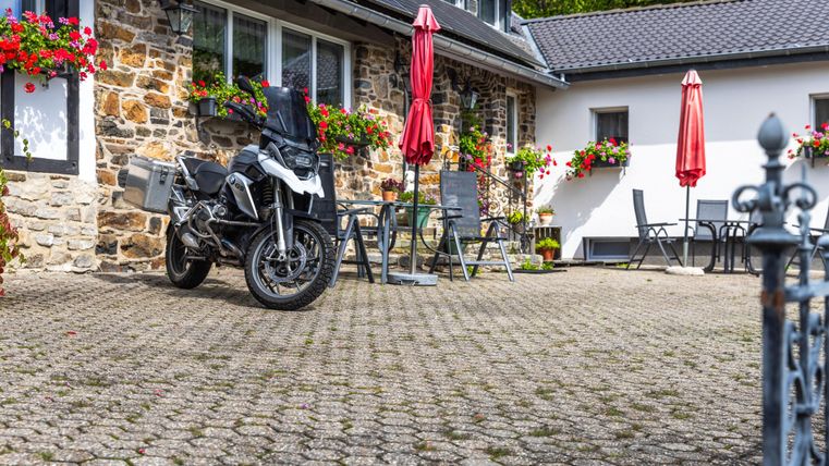 Een motorfiets staat op een geplaveide binnenplaats voor een mooi huis. Rode parasols en bloembakken geven de omgeving een uitnodigende sfeer.