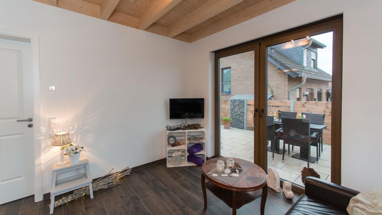 Een moderne woonkamer met houten balkenplafond en grote ramen. Er is een televisie, een tafel en toegang tot een buitenruimte.