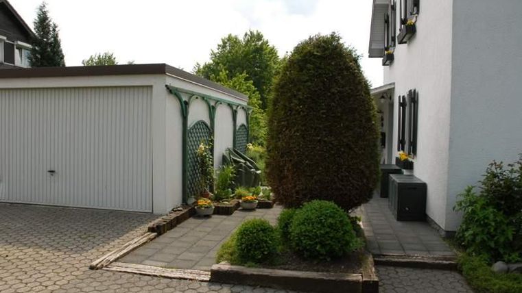 Een verzorgde entree met een haag en meerdere planten. Op de achtergrond is een eenvoudige aanbouw en een licht woongebouw te zien.