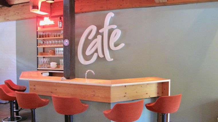 A modern café area with a wooden counter and orange chairs. The word "Café" is written on the wall in stylish letters.