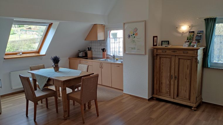 A bright kitchen nook with a dining table and chairs. In the background, the kitchen cabinets and a window with a view to the outside can be seen.