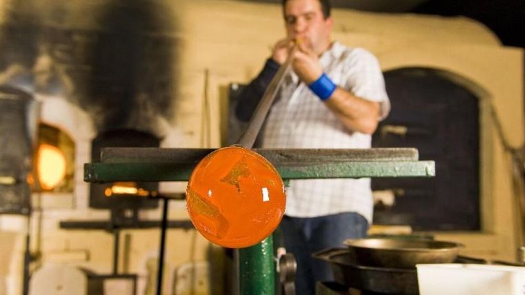 Een glasblazer werkt aan een glasstuk in een atelier. Op de achtergrond zijn ovens en gereedschappen te zien.