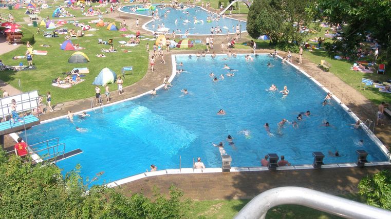 Een groot zwembad met veel zwemmers en recreatieve activiteiten. Er omheen zijn ligweiden en parasols geplaatst.