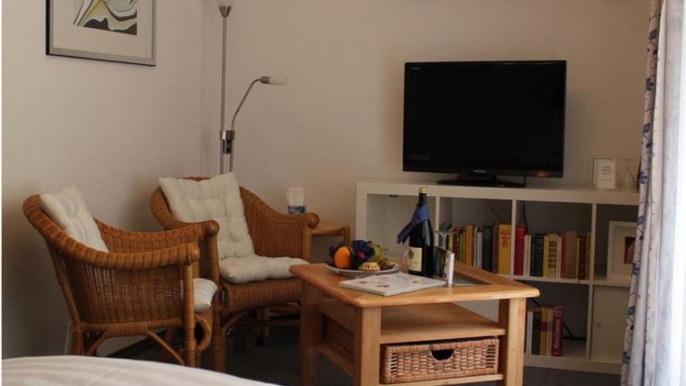 Ein gemütliches Wohnzimmer mit einem Fernseher und einem Couchtisch. Zwei Rattan-Stühle und Bücherregale sorgen für eine einladende Atmosphäre.