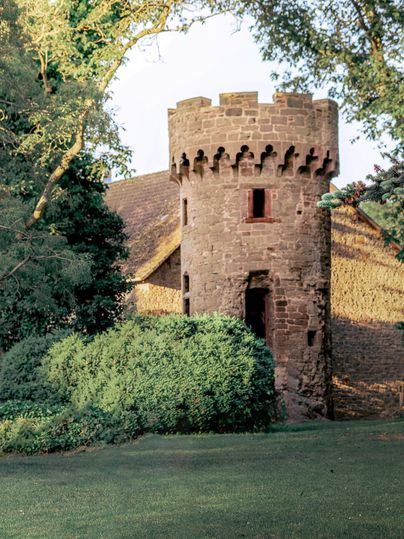Een kleine historische toren van steen omringd door groene struiken. Op de achtergrond is een oud gebouw en bomen zichtbaar.
