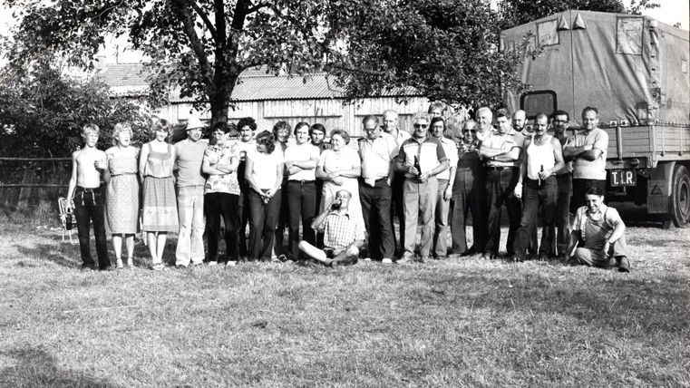A group of people stands in a meadow in front of a truck. They are smiling and posing together for a photo.