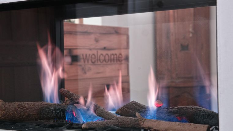 A modern fireplace with burning logs. In the background, there is a wooden box with the inscription "welcome."