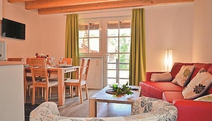 Ein gemütliches Wohnzimmer mit einem roten Sofa und einem Esstisch. Große Fenster mit grünen Vorhängen lassen viel Licht herein.