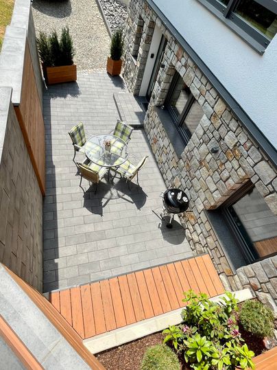 A modern terrace with a table and chairs. In the background, there is a stone building and a grill.