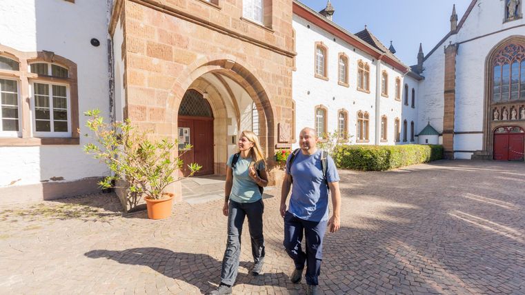 Zwei Personen spazieren in einem historischen Innenhof mit schönem Steinbau. Die Sonne scheint und es gibt grüne Pflanzen im Vordergrund.