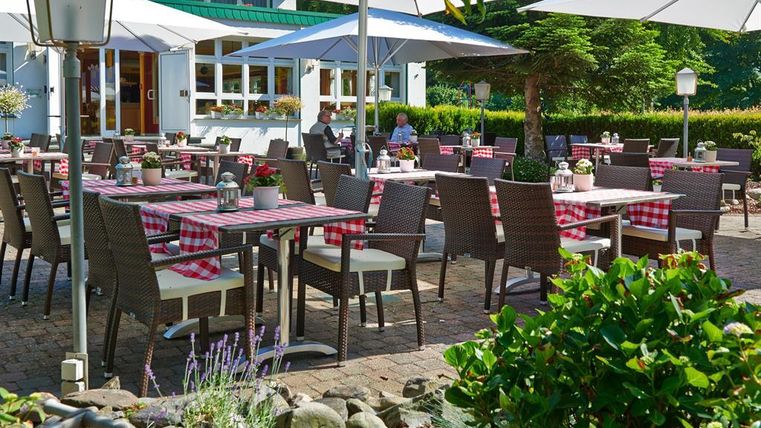 Ein einladendes Restaurant mit gemütlichen Tischen im Freien, dekoriert mit rot-weiß karierten Tischdecken. Umbrella bieten Schatten für die Gäste in einer ruhigen, grünen Umgebung.