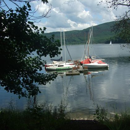 A calm lake with several boats gently floating on the water. Surrounded by green trees and hills under a cloudy sky.