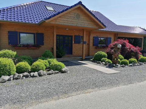 A cozy wooden house with a blue roof and green shrubs. The entrance is surrounded by colorful flowers and a well-maintained path.