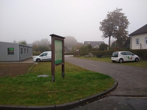 A foggy place with an information board and two cars in the parking lot. In the background, some buildings and trees are visible.