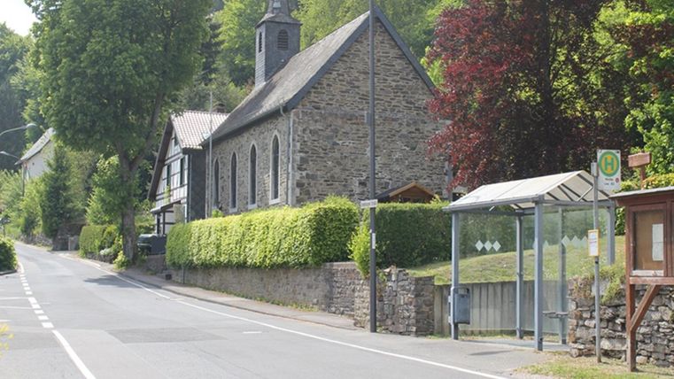 A quiet street with a stone church and green trees. There is a bus stop on the side.