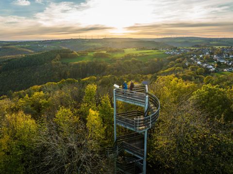 Een uitkijkplatform in de natuur met uitzicht op groene heuvels en een pittoresk dorp. De zon gaat achter de bergen onder.