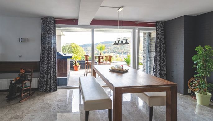 A modern dining room with a large wooden table and comfortable benches. Through the large windows, one looks out onto a green landscape.