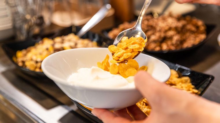 A person is filling a bowl with yogurt with cornflakes. In the background, various muesli and ingredients are visible in bowls.