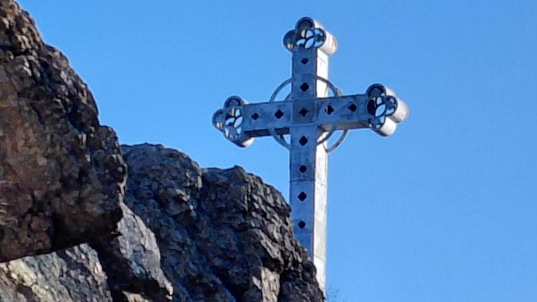 A large, silver cross stands on a rock under a clear blue sky. The surroundings are green and rocky.