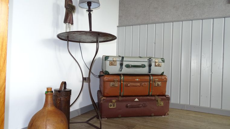 Een vintage tafel met een lamp staat naast een stapel oude koffers. Daaronder bevinden zich een grote fles en een kleine container.