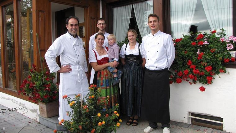A group of five people stands in front of a restaurant with blooming flowers. The people are wearing traditional costumes and chef uniforms.