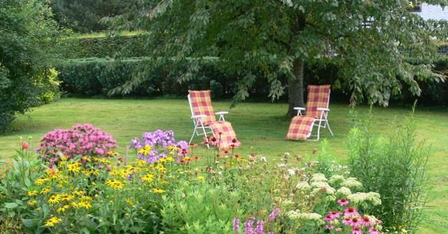 Een mooie tuin met kleurrijke bloemen en twee ligstoelen onder een boom. Het gras is groen en uitnodigend.