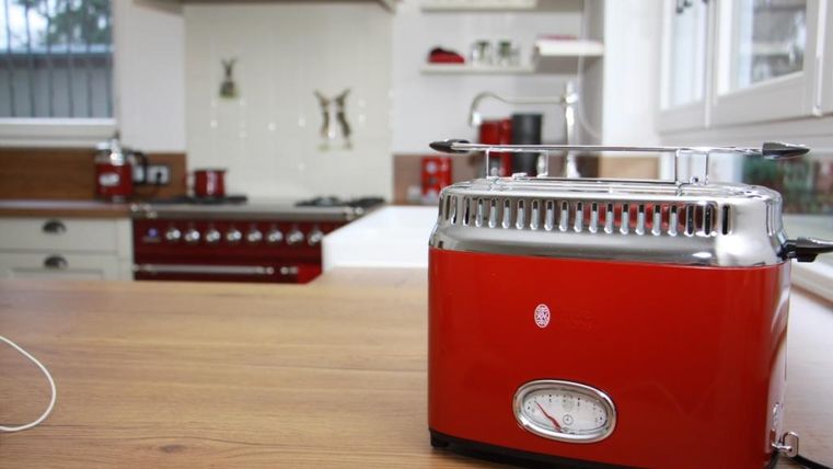A red toaster stands on a wooden board in a modern kitchen. In the background, a stove and shelves with dishes are visible.